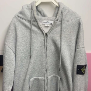 Grå hoodie från Stone Island - Säljer en grå hoodie från Stone Island med dragkedja, huva och snörning. Klassisk patch på ärmen och stora fickor framtill. Perfekt för dig som gillar streetwear och vill ha en clean look.