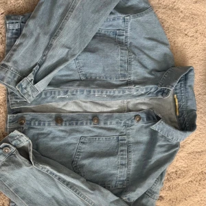 Ljusblå jeansjacka med knappar - Snygg ljusblå jeansjacka med klassisk krage och stora bröstfickor. Jackan har metallknappar framtill och på ärmsluten. Perfekt för dig som gillar en avslappnad och tidlös stil. Materialet är robust denim som ger en cool vibe.