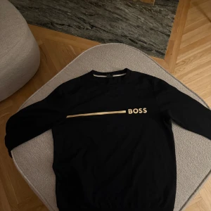 Svart sweatshirt från BOSS med guldtryck - Svart sweatshirt från BOSS med stilrent guldtryck och logga över bröstet. Tröjan har rund halsringning, långa ärmar och en enkel, clean design. Perfekt för dig som gillar en klassisk men ändå modern look.