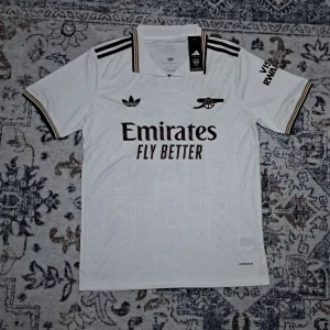 Arsenal vit fotbollströja Gyökeres #14 - Snygg Arsenal fotbollströja från Adidas i vit färg med svarta och guldiga detaljer. Kortärmad med klassisk krage, tryckta loggor och Gyökeres #14 på ryggen. Materialet är lätt och andas, Aeroready-teknologi för extra komfort. Perfekt för dig som vill ha en stilren och modern matchtröja.Arsenal 25/26 tredjeställ