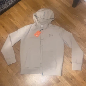 Beige hoodie från YoungLA med dragkedja - Snygg oanvänd beige hoodie från YoungLA med broderad logga på bröstet och huva med cool detalj. Tröjan har hel dragkedja framtill, två fickor och ribbade muddar vid ärmslut och nederkant. Perfekt för dig som gillar stilren streetwear.