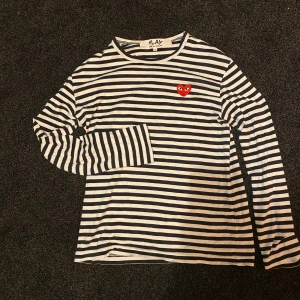 Randig långärmad tröja Comme des Garçons - Säljer en svartvit randig långärmad tröja från Comme des Garçons Play i storlek S, men den är liten i storleken. Tröjan är använd ytterst lite. Tröjan har ett ikoniskt rött hjärta med ögon broderat på bröstet och är gjord i mjuk bomull. 