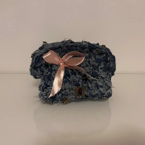 Virkad jeansväska med rosa rosett - Unik virkad handväska i blått denimtyg med fransig struktur och detaljer i brunt. Väskan har en söt rosa satinrosett framtill och en mjuk, rundad form. Perfekt för dig som gillar en kreativ och personlig stil.