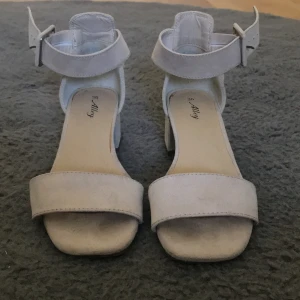 Beige sandaler med klack från Skopunkten - Snygga beige sandaler från Alley med bred rem över tårna och justerbart spänne runt ankeln. Skorna har låg klack och platt sula, tillverkade i mocka-liknande material. Perfekta för sommarens alla tillfällen. Nypris 300kr