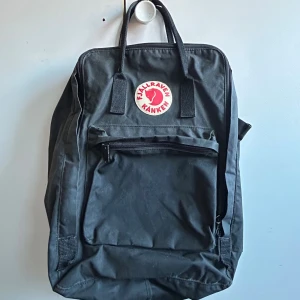 Svart Fjällräven Kånken ryggsäck - Fjällräven Kånken Laptop i färgen svart. Nypris 1799 kr. Jättesmidig och rymlig, säljer för att jag inte har någon användning för den. Ryggsäcken har ett stort huvudfack, en dragkedjeförsedd ytterficka, ett sidoficka, handtag och justerbara axelremmar.