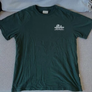 Mörkgrön Aimé Leon Dore t-shirt - Snygg mörkgrön t-shirt från Aimé Leon Dore med vit tryckt grafik och logga på både bröstet och ryggen. Klassisk passform med rund hals och korta ärmar. Tillverkad i mjuk bomull, perfekt för en clean och avslappnad stil. I nyskick! Nypris 800kr