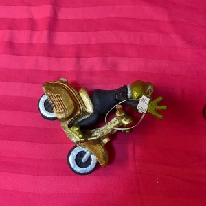 Groda på guldig scooter hängsmycke - Unikt hängsmycke föreställande en groda i svart kostym som kör en guldig scooter. Grodan har stora ögon och detaljer i grönt och vitt. Scootern är metallicguldig med svarta och vita hjul. Perfekt för dig som gillar roliga och annorlunda accessoarer.