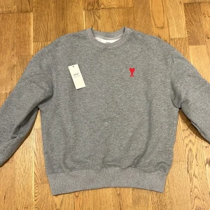 Grå sweatshirt från Ami - Säljer en snygg grå sweatshirt från Ami med ett rött hjärta broderat på bröstet. Tröjan är i nytt skick och har en klassisk rund halsringning. Perfekt för både höst och vår. Passar till alla tillfällen! Nypris 2800-3400