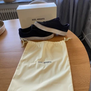 Axel Arigato svarta mocka sneakers - Snygga svarta sneakers från Axel Arigato i mocka med vit platt sula och elastiskt band över vristen. Klassisk låg modell med rund tå och diskret logga på sidan. Skorna levereras med originalpåse och box.