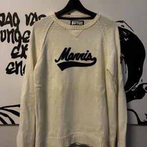 Vit stickad tröja från Morris - Vit stickad tröja från Morris med svart broderad logga på bröstet och Union Jack-detalj på ärmen. Tröjan har rund halsringning, ribbade muddar och lång ärm. Perfekt för dig som gillar klassisk och sportig stil.