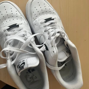 Nike Air Force 1 vita sneakers - Säljer ett par klassiska Nike Air Force 1 sneakers i vitt skinn med svart swoosh. Skorna har perforerad tå, vit platt sula och snörning. Perfekta för dig som vill ha en clean och tidlös look med streetkänsla.