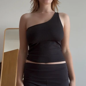 One shoulder topp  - Säljer en svart ribbad one shoulder topp från Gina Tricot i storlek D. Toppen är tight och har en asymmetrisk axelbandslös design med smal axelrem på ena sidan. 