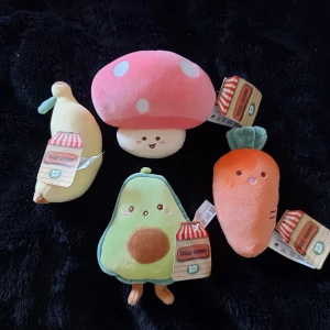 Huggie Greenies Plush Set - Fyra mjuka och färgglada gosedjur i form av svamp, banan, avokado och morot. Alla med söta ansikten och etiketter kvar. Perfekt skick, inga synliga defekter eller slitage. Passar som present eller dekoration.
