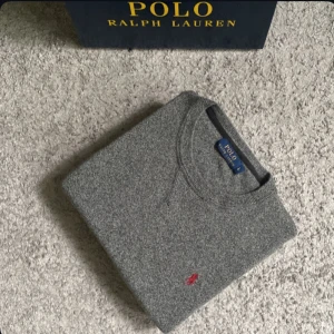 Grå stickad tröja Polo Ralph Lauren - Snygg grå stickad tröja från Polo Ralph Lauren med klassisk rund halsringning och röd broderad logga på bröstet. Tröjan har ribbade muddar vid ärmslut och nederkant. Lappen där bak är lite av 