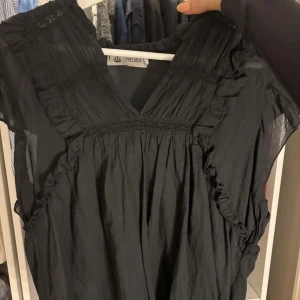 Svart blus från Chelsea - Supersöt svart blus från Chelsea med v-ringning och volangdetaljer längs axlar och bröst. Tunn, luftig bomullskvalitet och korta ärmar. Perfekt för dig som gillar en romantisk och avslappnad stil.