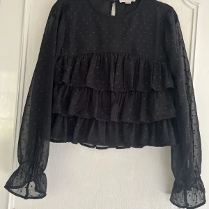 Svart volangblus från Gina Tricot - Supersöt svart blus från Gina Tricot med flera volanglager och små glittriga prickar. Blusen har transparenta långärmade ärmar med resår och volang vid ärmslut. Perfekt för dig som gillar en trendig och lekfull stil💕😻 Säljer för att jag har vuxit ur denna