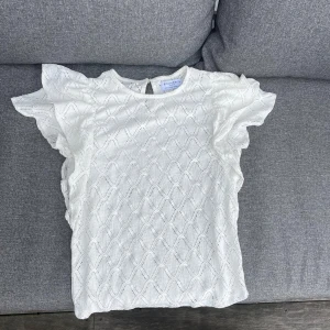 Vit blus med volangärmar Holly & White - Säljer en söt vit blus från Holly & White med volangärmar och diskret strukturmönster. Blusen har rund halsringning och knäppning bak i nacken. Perfekt för dig som gillar romantiska detaljer och en luftig känsla.