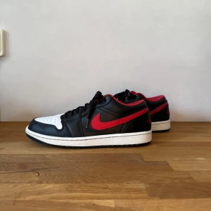 Nike Air Jordan 1 Low svart/röd/vit - Säljer ett par Nike Air Jordan 1 Low med svart och vitt skinn, röda detaljer och klassisk röd swoosh. Skorna har svart snörning, perforerad tåbox och röd insida. Perfekta för dig som vill ha en ikonisk sneaker med snygg färgkombo.