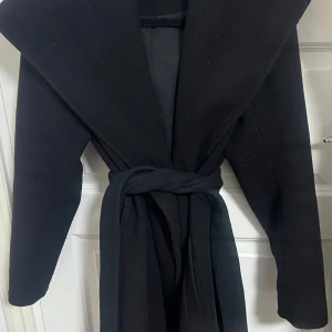 Svart kappa med bälte och stor krage - Stilren svart kappa med bred krage och knytskärp i midjan. Jackan har lång ärm och en snygg, rak passform. Perfekt för dig som vill ha en klassisk look med modern twist. Materialet är mjukt och känns lyxigt, och kappan har fickor i sidorna. Kan passa M storlek med. 