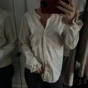Vit volangskjorta från Mos Mosh - Säljer en stilren vit skjorta från Mos Mosh med volangdetaljer vid ärmsluten och v-ringad hals. Skjortan har lång ärm och knappar framtill. Som en storlek S/M