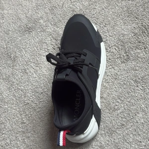 Svarta sneakers från Moncler - Snygga svarta sneakers från Moncler med vit sula och svarta snören. Skorna har Monclers klassiska röd-vit-blå detalj baktill och en unik mönstrad yttersula. Kvitto och box och Monclear påse finns, skriv om mer info!!