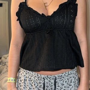 Svart blus med broderade detaljer - Säljer en svart blus med tunna axelband och volangkant upptill. Toppen har broderade hålmönster och en lätt utsvängd passform. Perfekt att styla med jeans eller kjol för en trendig look.