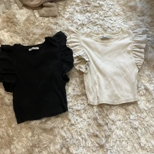 2 toppar med volangärmar från Zara - Två snygga toppar från Zara med volangärmar, en i svart och en i beige. Båda har rund halsringning och är tillverkade i mjuk bomull. Perfekta för att lyfta din outfit med en cool detalj på axlarna.