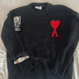 Svart stickad tröja från Ami Paris - Svart stickad tröja från Ami Paris med rund halsringning och ribbade muddar. På bröstet finns ett rött hjärta med bokstaven A, som är typiskt för märket. Tröjan är i mjuk ull och har en klassisk, avslappnad passform. Perfekt för dig som gillar stilrena detaljer.