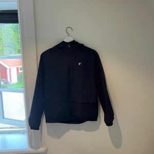 Svart vindjacka från Lyle & Scott - Svart vindjacka från Lyle & Scott med huva och dragkedja framtill. Jackan har två stora fickor med knappar och den klassiska gula loggan på bröstet. Perfekt för blåsiga dagar och enkel att matcha med allt.