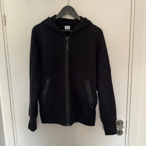 Svart zip hoodie från C.P. Company - Svart hoodie från C.P. Company med dragkedja framtill, två fickor med dragkedja och en extra ficka på ärmen. Klassisk huva och långärmad modell, perfekt för en stilren och avslappnad look.