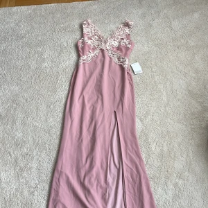 Rosa långklänning med spetsdetaljer - Supersnygg rosa långklänning från Moments New York med v-ringning fram och i ryggen, spetsdetaljer och blommigt broderi upptill. Klänningen har en slits framtill och är ärmlös, perfekt för speciella tillfällen. Materialet känns mjukt och följsamt. Aldrig använd med prislapp kvar