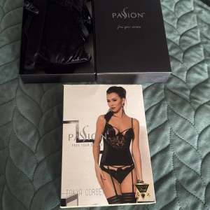Svart korsett med spets från Passion - Säljer en svart korsett från Passion, modell Tonya Corset. Den har justerbara axelband, bygel och hakknäppning bak. Materialet är glansigt med vackra spetsdetaljer och blommigt mönster framtill. Perfekt för dig som vill ha en snygg och feminin look.