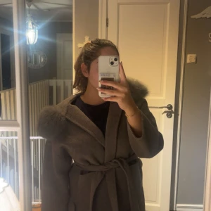 Grå kappa med pälskantad huva - Säljer en Grå kappa med stor fluffig pälskantad huva och knytskärp i midjan. Jackan har en loose fit och är tillverkad i ullblandning, perfekt för kyliga dagar. Stilen är enkel men ändå lyxig med breda ärmar och markerad midja.