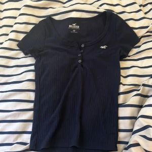 Mörkblå ribbad topp från Hollister XS - Säljer en mörkblå ribbad topp från Hollister Must Have Collection i storlek XS. Toppen har korta ärmar, rund halsringning och knappar framtill. Liten broderad Hollister-logga på bröstet (defekt). Perfekt basic med snygg passform.