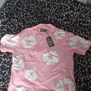 Rosa blommig t-shirt från Denim Tears - Säljer en rosa t-shirt från Denim Tears med stora vita blommor tryckta över hela tröjan. Klassisk passform med rund hals och korta ärmar. Materialet är mjuk bomull och designen är riktigt unik och sticker ut.