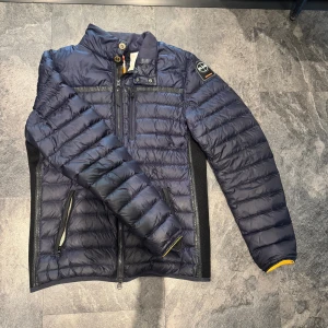 Mörkblå pufferjacka Parajumpers - Snygg mörkblå pufferjacka från Parajumpers med quiltad design, hög krage och dragkedja framtill. Jackan har svarta detaljer på sidorna, två fickor med dragkedja och en ikonisk patch på ärmen. Insidan är ljusbeige med orange detaljer. !! Byte går också !!