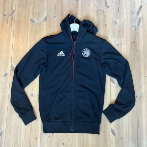 Svart Adidas hoodie Ajax med dragkedja - Svart hoodie från Adidas med Ajax-logga på bröstet och vit Adidas-logga. Tröjan har huva med snörning, hel dragkedja och två fickor framtill. Perfekt för dig som gillar sportig stil och vill representera Ajax.