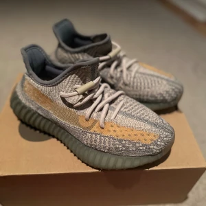 Adidas Yeezy Boost 350 V2 Ash Stone - Säljer ett par Adidas Yeezy Boost 350 V2 Ash Stone med stickad ovandel i grått och beige, samt en orange-brun stripe på sidan. Skorna har en chunky, ribbad sula i mörkgrönt och klassisk snörning. Perfekta för dig som vill sticka ut med en unik sneaker.