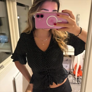 Svart prickig blus med volangärm - Säljer en svart blus med vita prickar och v-ringning. Blusen har korta volangärmar och rynkade detaljer framtill samt vågig kant nertill. Perfekt för dig som gillar en söt och trendig look.