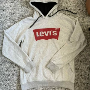 Grå hoodie från Levi's med logga - Säljer en grå hoodie från Levi's med stor röd logga framtill. Tröjan har huva med svarta snören, magficka och ribbade muddar vid ärmslut och nederkant. Perfekt för en avslappnad och cool stil.