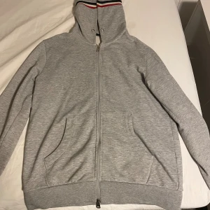 Grå Moncler hoodie med dragkedja - Grå hoodie från Moncler med hel dragkedja, huva med röd, vit och marinblå rand samt två stora fickor framtill. Klassisk Moncler-logga på ärmen och ribbade muddar. Perfekt för en chill och stilren look.