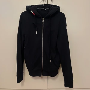 Marinblå kofta från Moncler med dragkedja - Marinblå kofta från Moncler med hel dragkedja framtill, huva med snörning och klassisk Moncler-logga på ärmen. Tillverkad i mjuk bomull och har ribbade muddar. Mannen fick denna i present för ett par år sedan och denna ska vara äkta och inköpt på Pandorf. Storlek XS, mycket fint skick och inga defekter. 