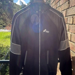 Asics retro track top storlek M - Sportig svart vindjacka från Asics med vita och grå detaljer. Jackan har dragkedja framtill, prickiga partier på axlar och ärmar samt Asics-logga på bröstet och längs ärmarna. Perfekt för dig som gillar en clean och atletisk look. Bra skick, inga skavanker