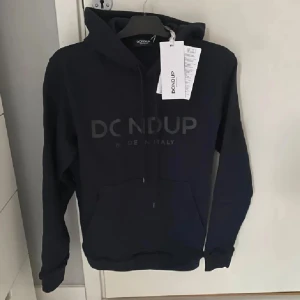 Svart hoodie från Dondup med huva - Svart hoodie från Dondup med stor logga framtill. Tröjan har huva med snörning och en klassisk känguruficka. Tillverkad i mjuk bomull och har långa ärmar. Perfekt för en avslappnad och stilren look. Lapparna finns inte kvar!