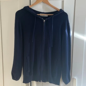 Kashmir zip marinblå  - Mörkblå hoodie från Zadolo med dragkedja framtill, huva med snörning och två fickor. Tröjan är stickad och har lång ärm, perfekt för kyligare dagar. Enkel och stilren design som funkar till det mesta.