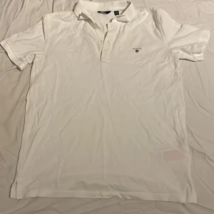 Vit pikétröja från Gant - Klassisk vit pikétröja från Gant med korta ärmar och liten broderad logga på bröstet. Tröjan har krage och knappar framtill, perfekt för en clean och stilren look. Tillverkad i mjuk bomull som känns skön mot huden. 