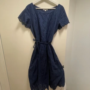 Marinblå klänning Hampton Republic - Snygg marinblå klänning från Hampton Republic i storlek 40. Klänningen har korta ärmar med broderade spetsdetaljer, knytband i midjan och ett vackert hålmönster över hela plagget. Tillverkad i mjuk bomull, perfekt för dig som gillar en feminin och bekväm stil.