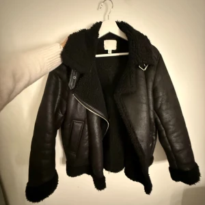 Svart pälsjacka med dragkedja - Säljer en svart pälsjacka från H&M med bred krage och fluffiga detaljer vid ärmslut och nederkant. Jackan har en snygg asymmetrisk dragkedja och två fickor framtill. Perfekt för dig som gillar en cool och varm look. Passar S och M