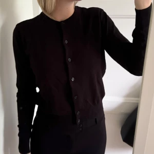 Svart kofta från H&M med knappar - Svart klassisk kofta från H&M med rund hals och knappar hela vägen framtill. Långärmad och tillverkad i mjuk bomullsmix. 