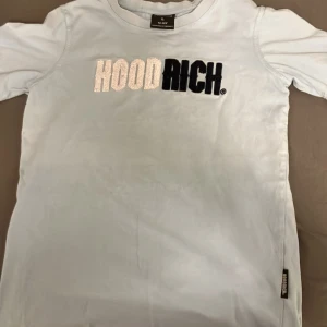 Ljus blå HOODRICH t-shirt - Snygg ljusblå t-shirt från HOODRICH med stort broderat logotyp-tryck i vitt och svart på bröstet. Klassisk passform med korta ärmar och rund hals. Perfekt för en streetwear-look och enkel att matcha med jeans eller shorts.